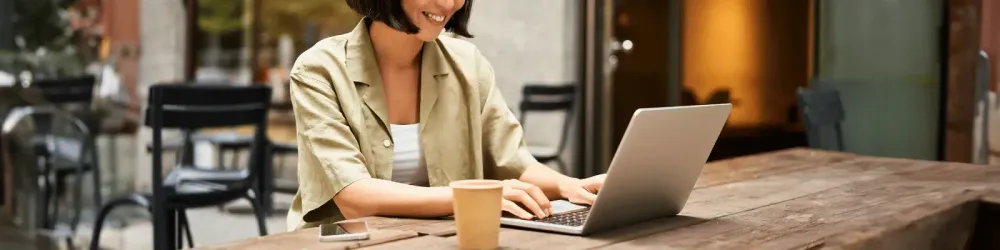 Young asian woman, digital nomad working remotely from a cafe, drinking coffee and using laptop, smiling